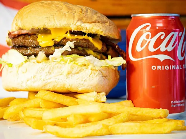 A stacked smash burger with melted cheese and crispy bacon, served with golden fries and a can of Coca-Cola.